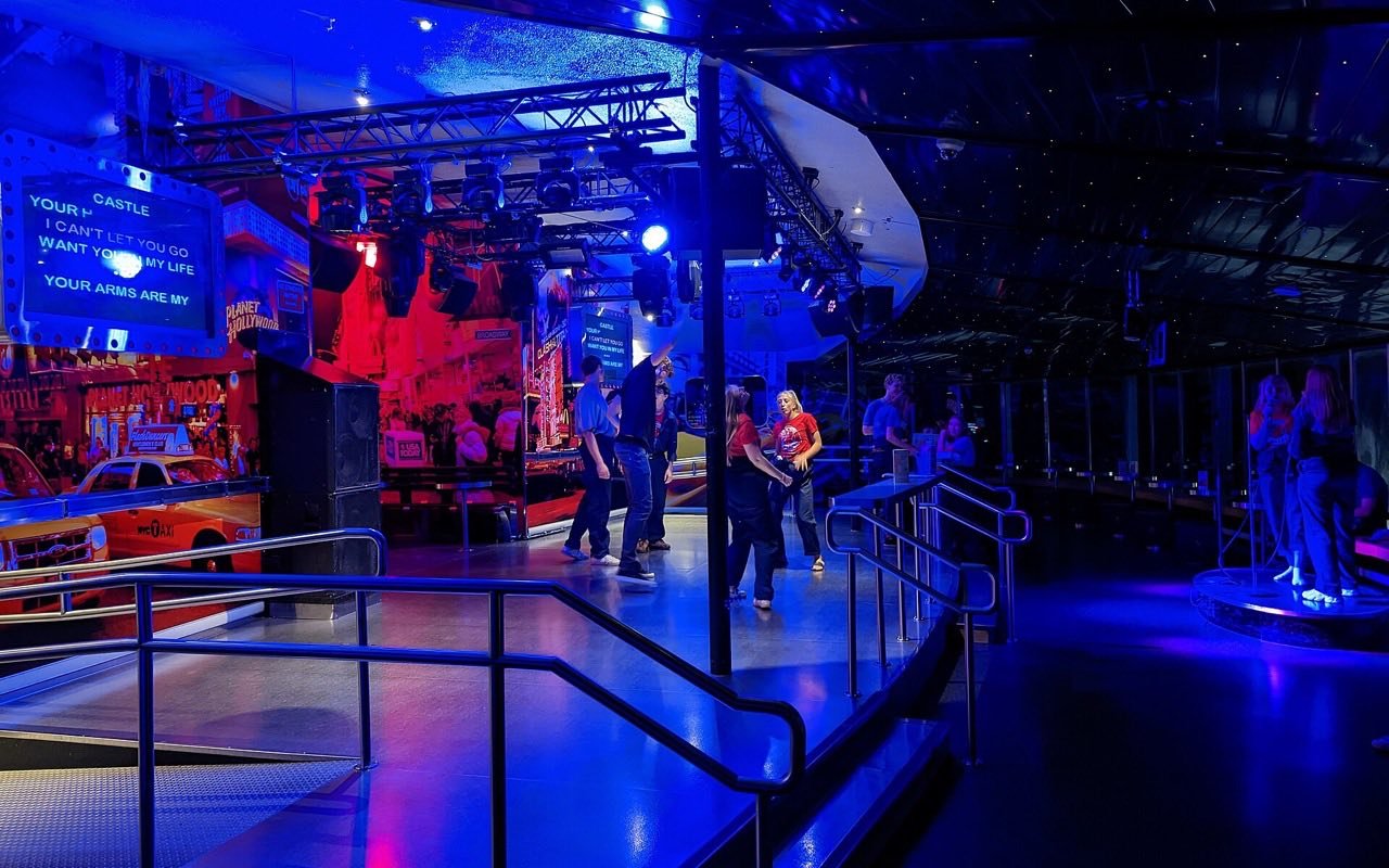 Symbolbild Karaoke: Gäste auf einer Clubbühne in blauem und rotem Licht, links ein Bildschirm mit Text