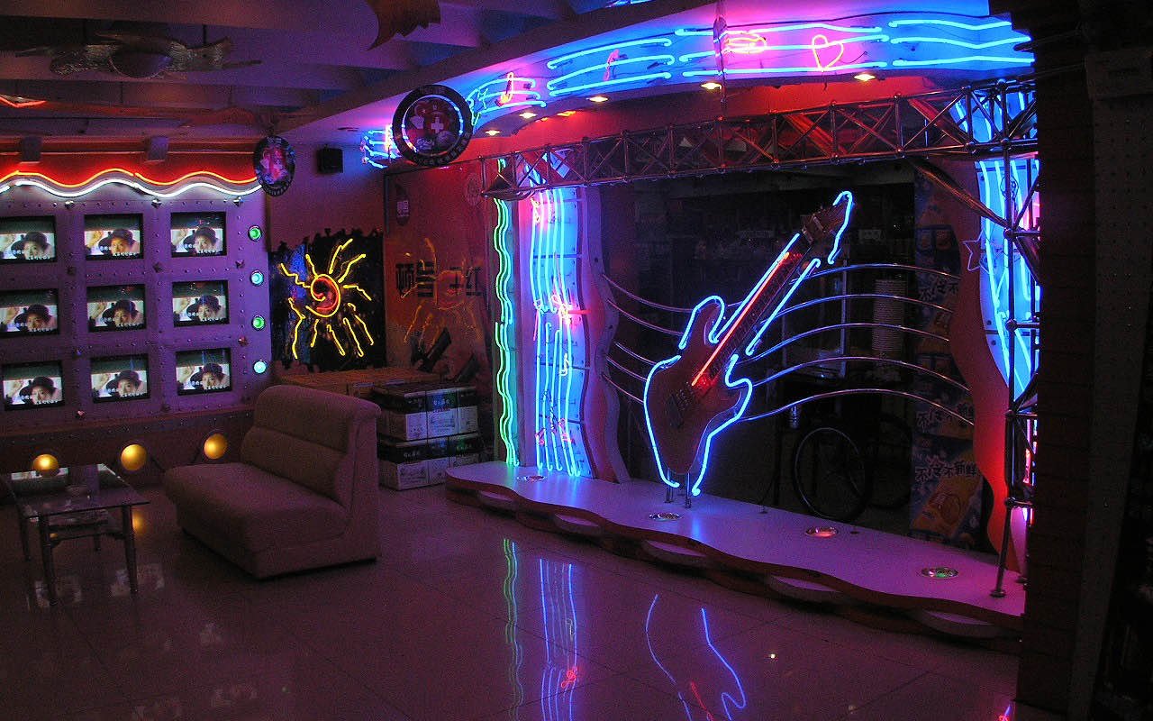 Symbolbild Karaoke: Kleine Bühne mit Gitarre aus Neonlicht, Sofa und Monitorwand in einem Karaokelokal